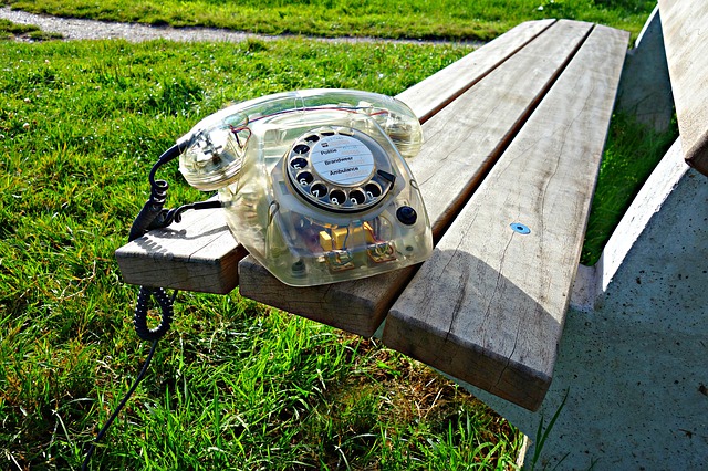 Telefoon met draaischijf, doorschijnend plastic, op een houten bankje in het park. Zoek je snel contact? Bellen werkt het snelste!